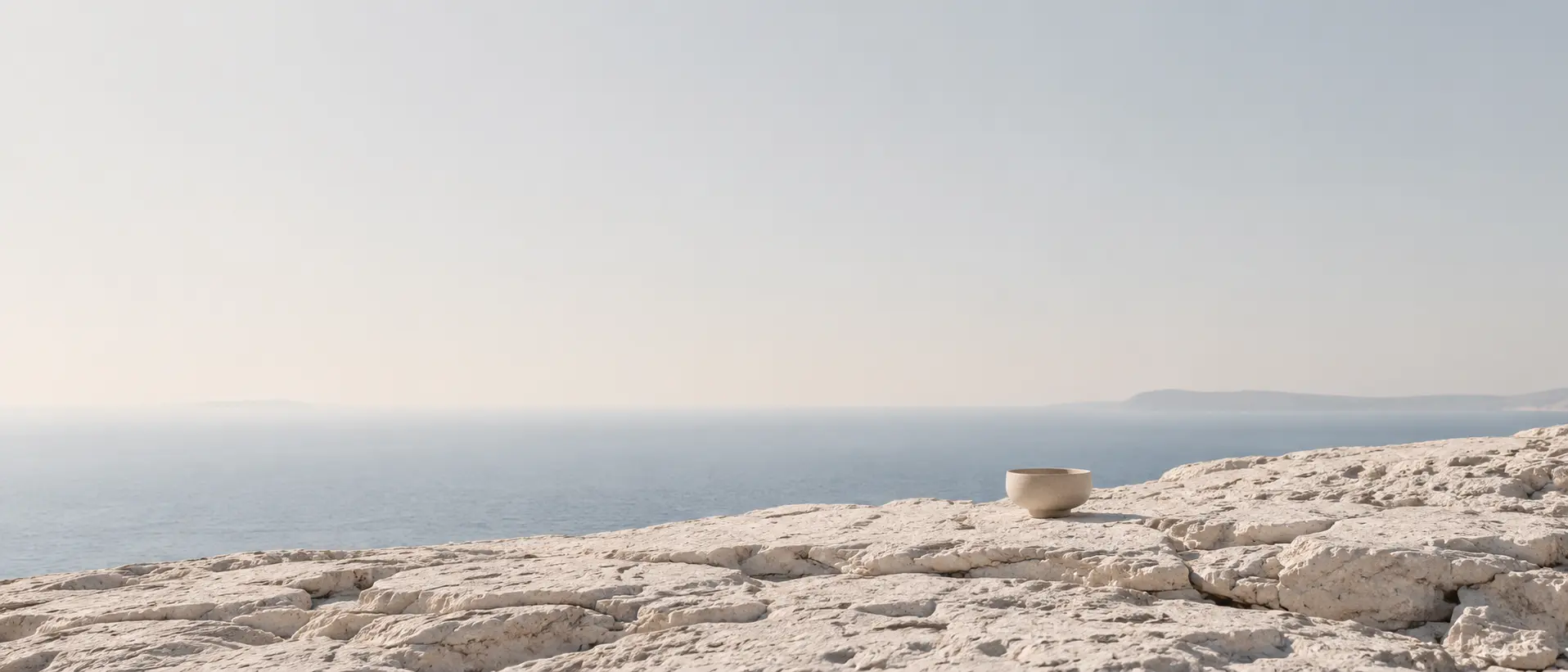 A ceramic vessel standing on a limestone rock shelf above the Aegean sea, in soft Cycladic light