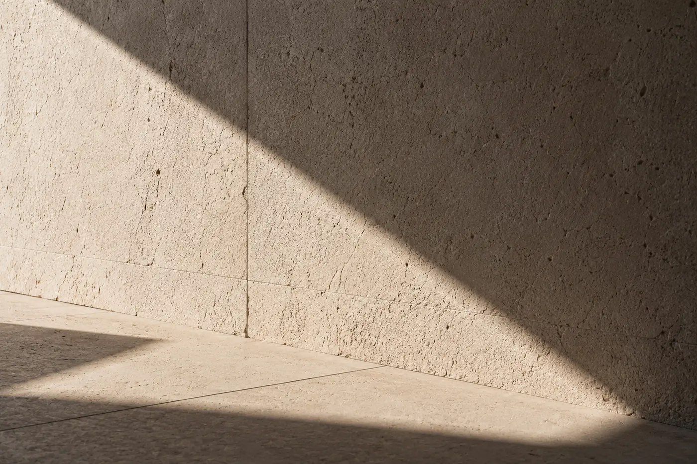 A sharp diagonal of sunlight crossing a porous travertine wall and stone floor